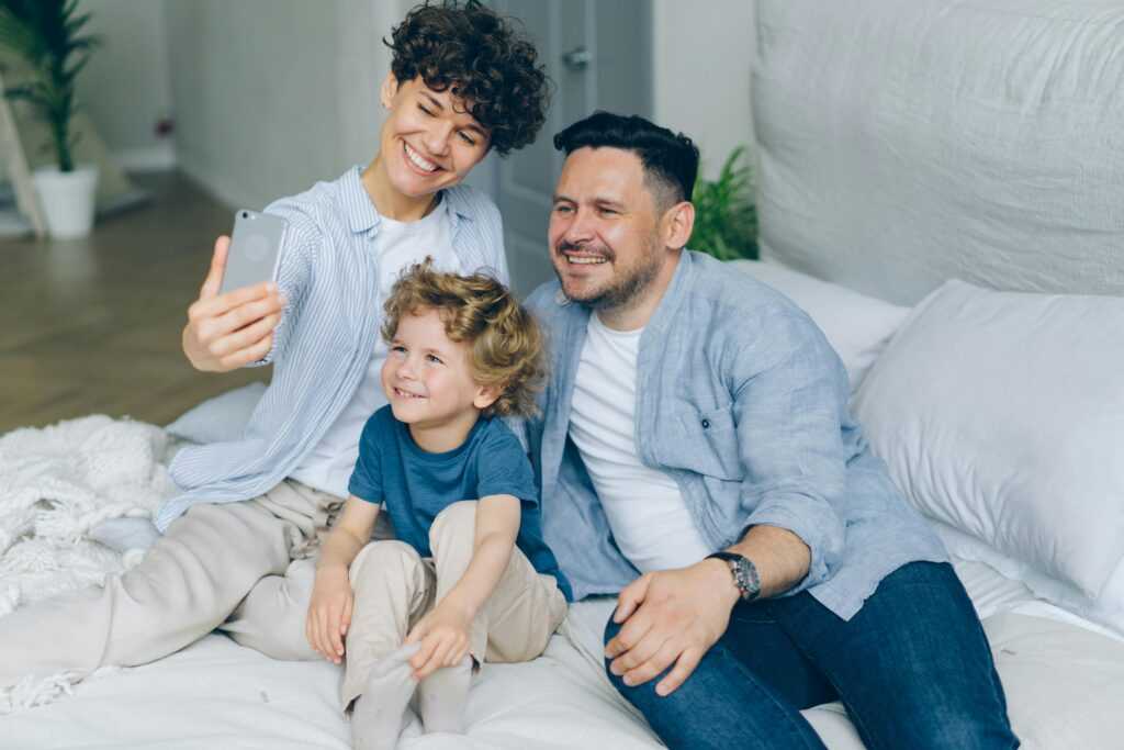 Smiling parents and child taking a selfie on a bed, capturing a joyful moment together.